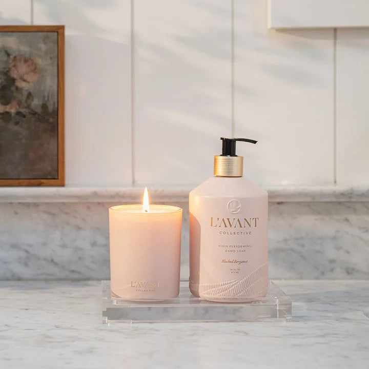 Lit L’AVANT Collective Blushed Bergamot candle displayed beside matching hand soap on a clear vanity tray.