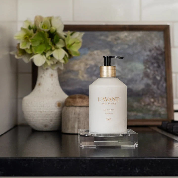 L’AVANT Collective Blushed Bergamot Hand Lotion displayed on a clear acrylic tray with flowers and neutral kitchen decor