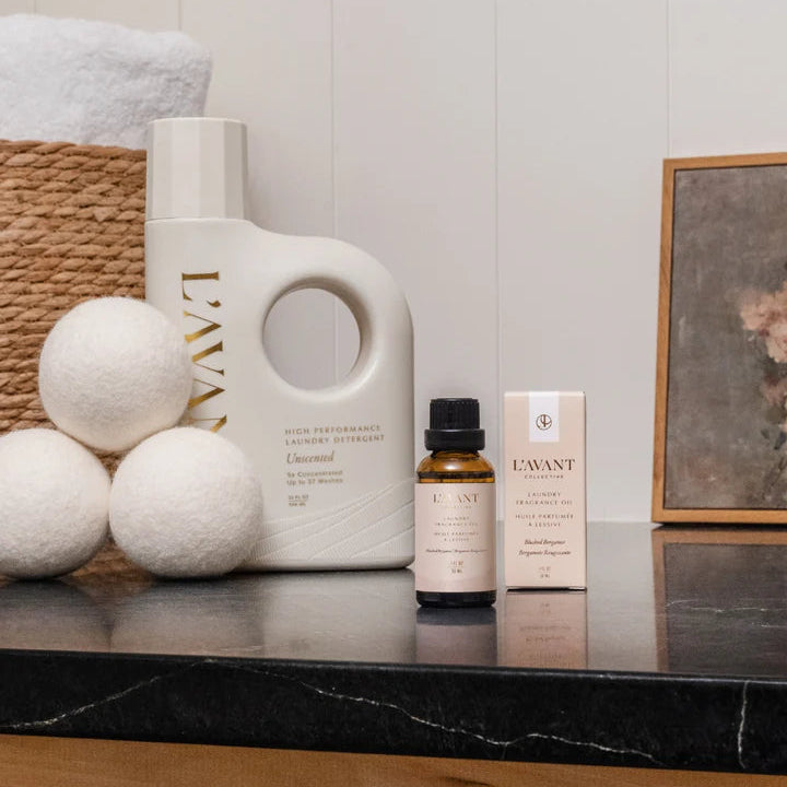 L’AVANT Collective Blushed Bergamot Laundry Fragrance Oil displayed with unscented detergent and white wool dryer balls.