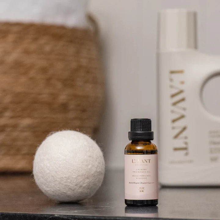 L’AVANT Collective Blushed Bergamot Laundry Fragrance Oil displayed beside a wool dryer ball and laundry detergent.