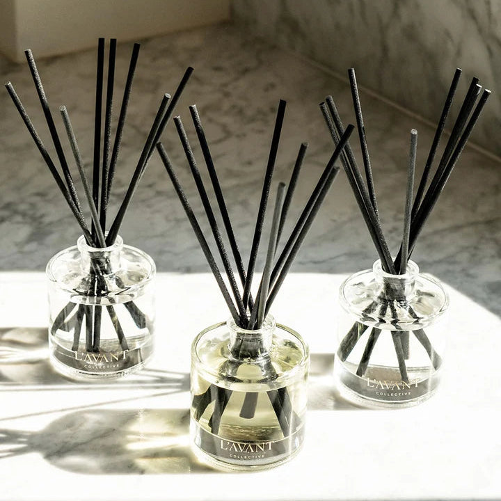 Three L’AVANT Collective glass reed diffusers with black reeds styled together in natural sunlight.