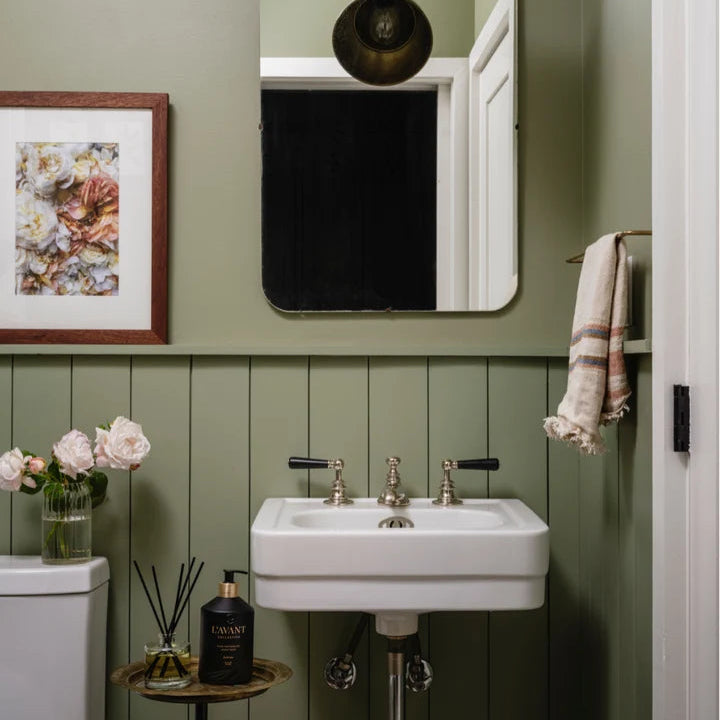 L’AVANT Collective Fresh Linen reed diffuser and hand soap styled beside a white sink in a green bathroom.