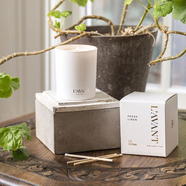 L’AVANT Collective Fresh Linen scented candle and product box styled with greenery on a rustic wood table.