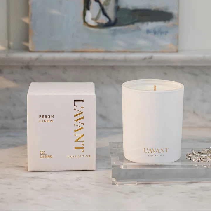 L’AVANT Collective Fresh Linen candle displayed with its white and gold product box on a marble countertop.