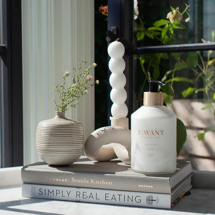 L’AVANT Collective Fresh Linen Hand Lotion styled on stacked books with neutral home decor near a window