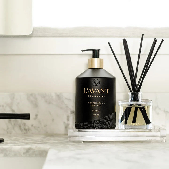L’AVANT Collective Fresh Linen reed diffuser and black hand soap displayed together on a clear countertop tray.