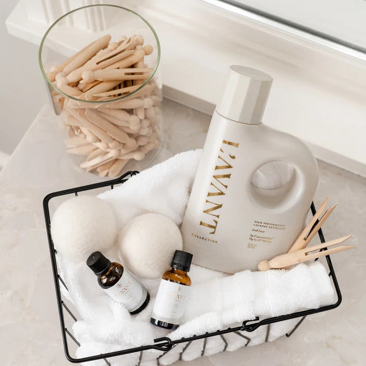L’AVANT Collective Laundry Fragrance Oil and detergent arranged in a laundry basket with wool dryer balls, towels, and clothespins.