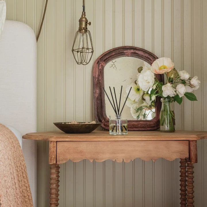 L’AVANT Collective reed diffuser with black reeds styled on a wooden bedside table with flowers and vintage mirror.