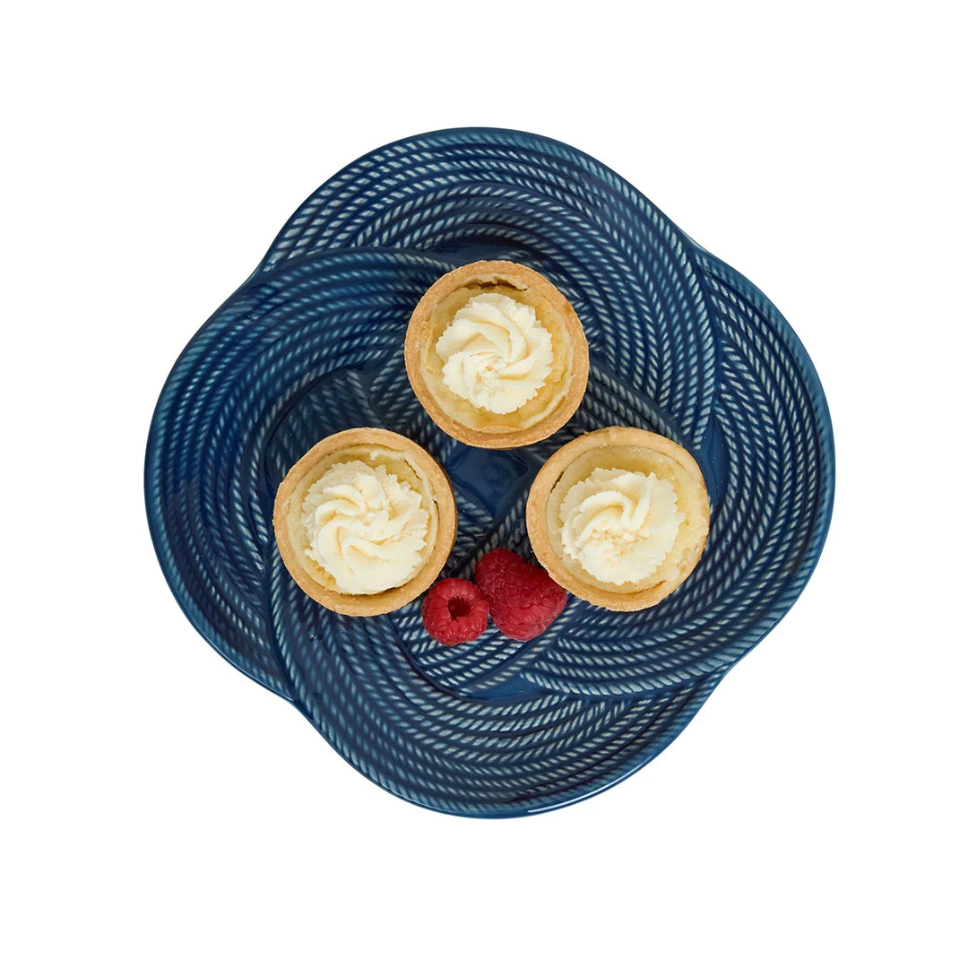 Angled view of the Le Panier Mystic Knot Dessert/Salad Plate in Denim showcasing its woven rim and sculptural blue knot detailing.