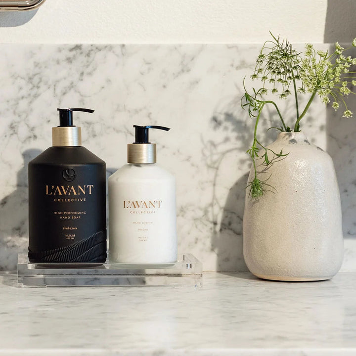 Clear lucite countertop tray holding hand soap and hand lotion beside a decorative vase on a marble counter.