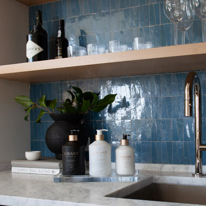 Clear lucite kitchen sink tray holding soap and lotion dispensers on a marble countertop against a blue tile backsplash.
