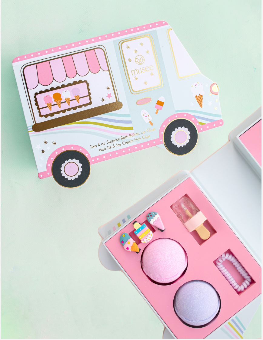 Musee ice cream truck bath balm and accessory set shown with the truck package and open box containing two bath balms, ice cream hair clips, lip gloss, and a hair tie.