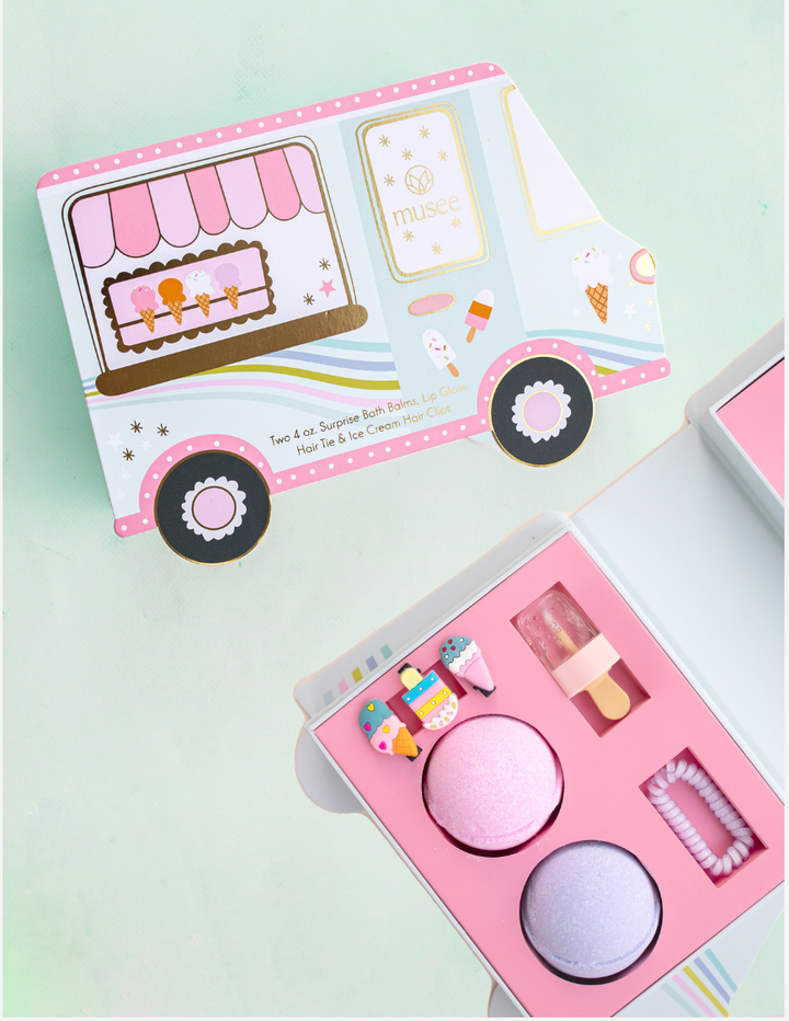 Musee ice cream truck bath balm and accessory set shown with the truck package and open box containing two bath balms, ice cream hair clips, lip gloss, and a hair tie.