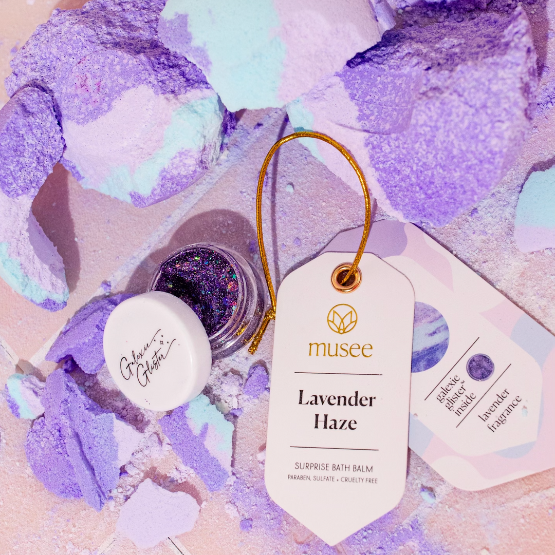 Musee Lavender Haze surprise bath balm broken with its surprise body/hair glitter revealed, displayed next to its product tag. 