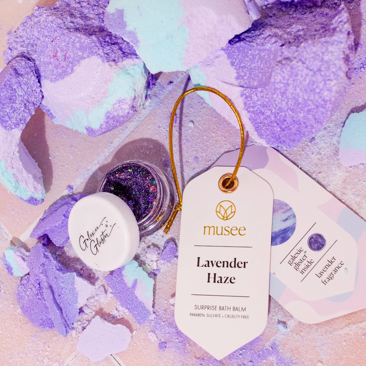 Musee Lavender Haze surprise bath balm broken with its surprise body/hair glitter revealed, displayed next to its product tag. 