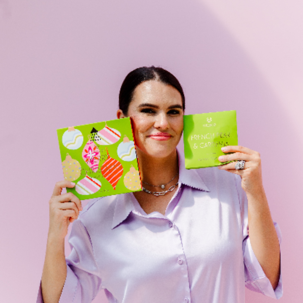 Woman holding Musee's Merry Christmas Shower Steamer Set and Bath Salts, while smiling in front of a purple wall. 