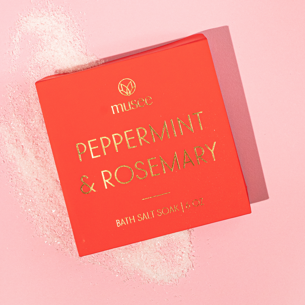 Musee's Peppermint & Rosemary Bath Salt Soak scattered across a pink background underneath its packaging. 