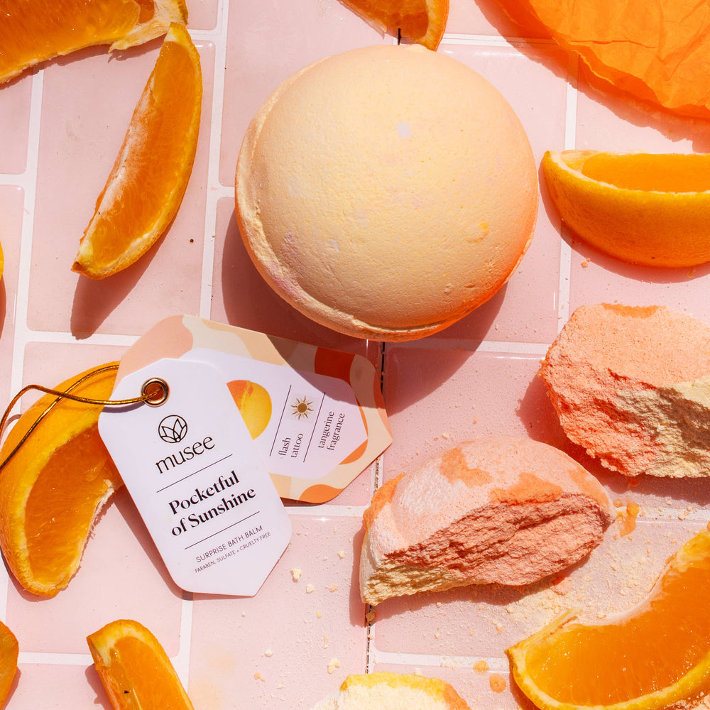 Musee Pocketful of Sunshine bath balm surrounded by fresh orange slices and  bath balm pieces.