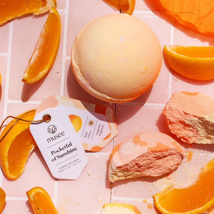 Musee Pocketful of Sunshine bath balm surrounded by fresh orange slices and  bath balm pieces.