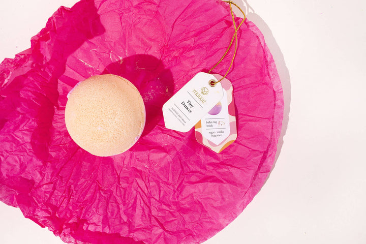 Musee Tiny Dancer surprise bath balm unwrapped and rested on its bright pink tissue paper next to its decorative product tag.