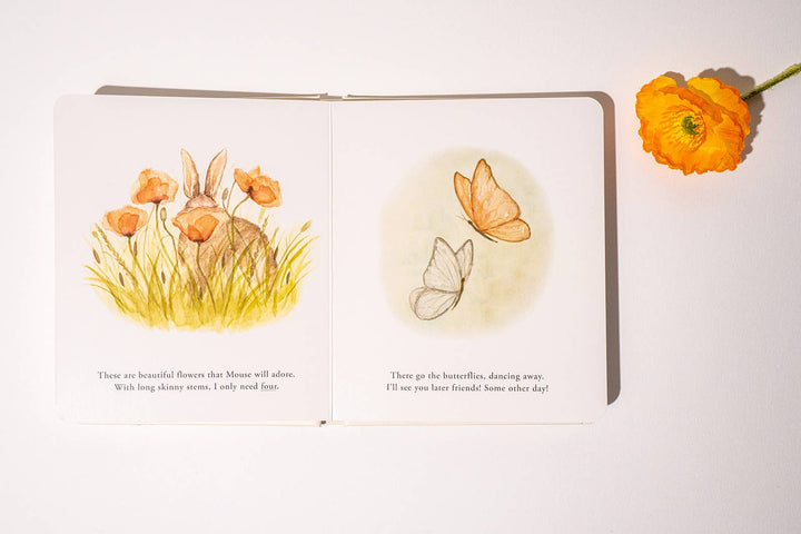 Open Our Little Adventures board book showing watercolor flowers and butterflies with an orange flower beside the book