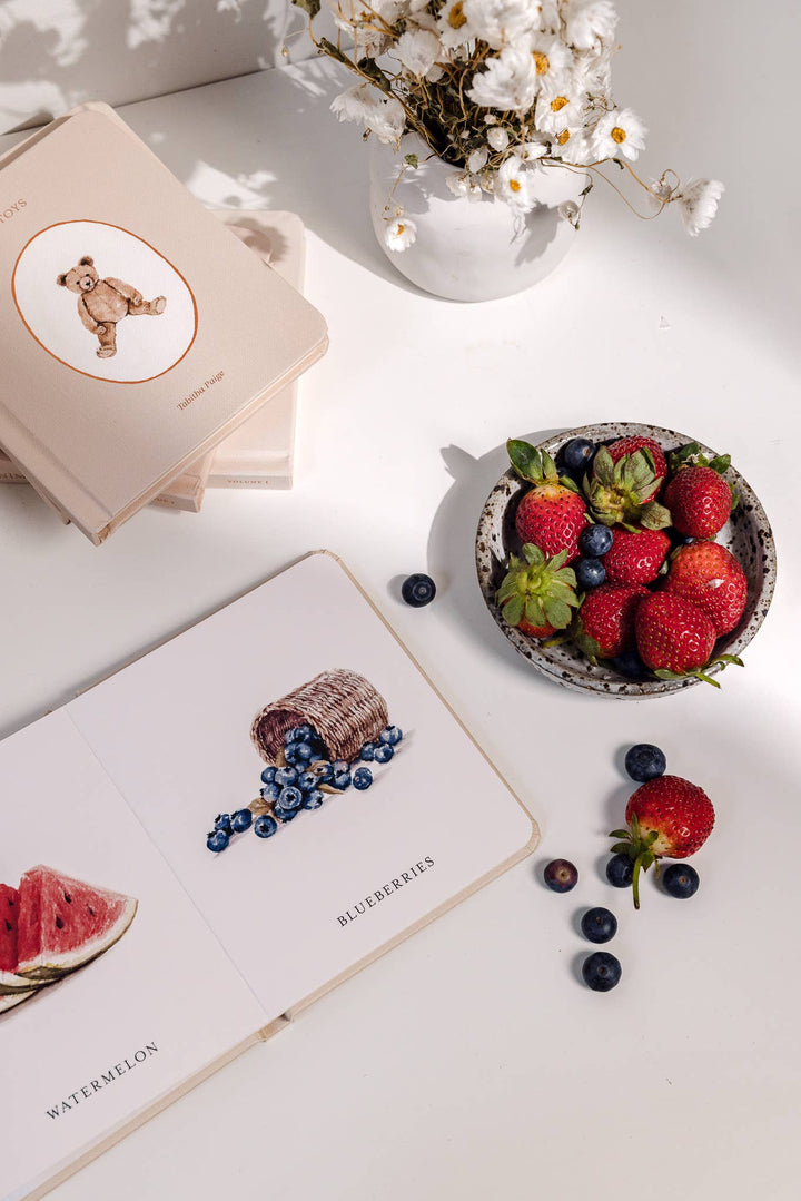 Our Little Library Vol. 1 board book opened to illustrated blueberries and watermelon vocabulary pages beside fresh berries