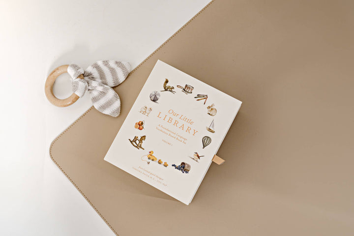 Our Little Library Vol. 1 five board book box set styled on a neutral mat beside a wooden baby teether