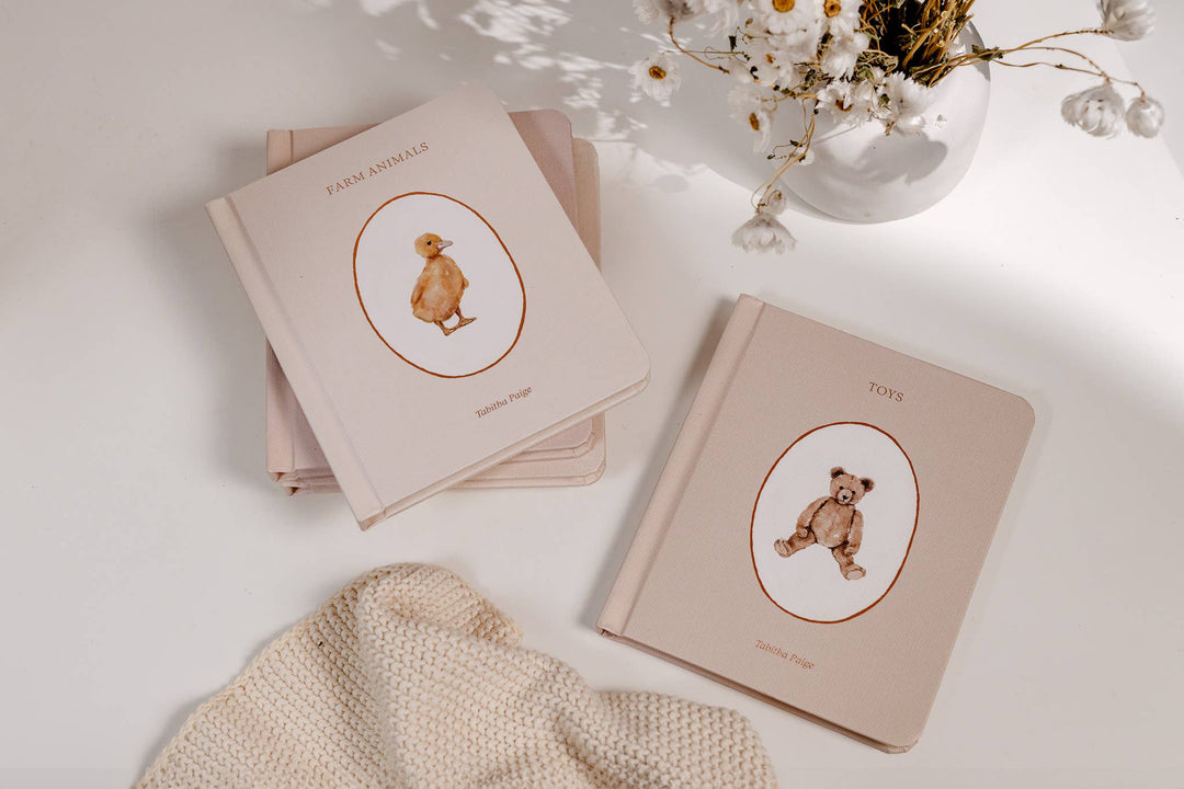 Our Little Library Vol. 1 board books styled with Farm Animals and Toys covers beside a vase of flowers and knit blanket