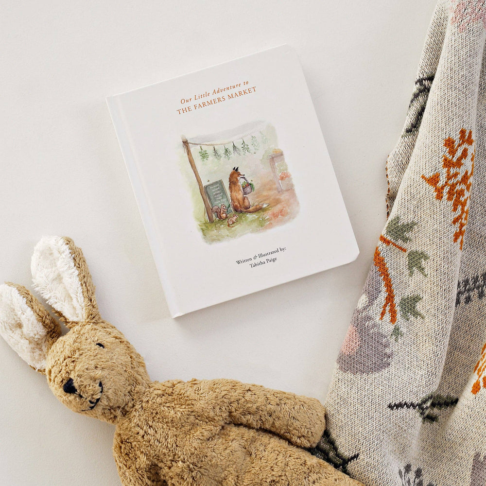 Styled Image of Paige Tate Co.'s Our little Adventure to the Farmers Market book laying next to a blanket and a stuffed brown bunny toy. 