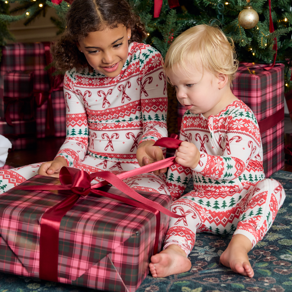 Lifestyle image of a little girl wearing Pink Chicken's Kids Bamboo PJ Set and an infant wearing the Baby Bamboo Romper, both in their new 'Candy Cane Fair Isle' Print. 