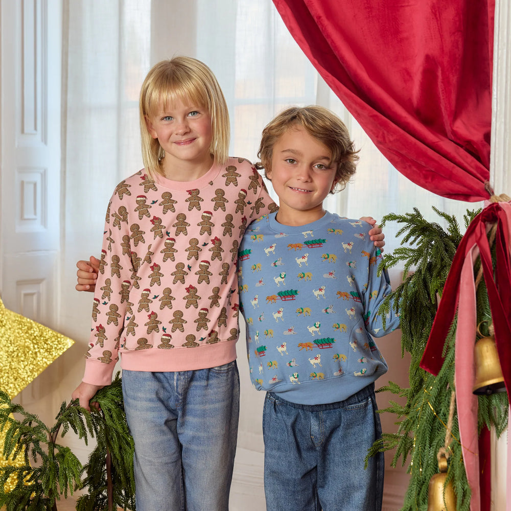 Young boy wearing the blue Holiday Labs Play Sweatshirt with jeans, standing beside a girl in a pink gingerbread sweatshirt for Christmas.