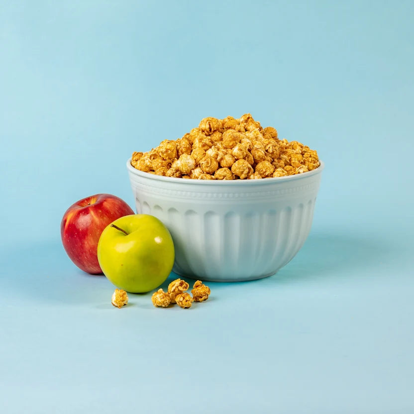 Bowl filled with golden caramel apple popcorn alongside fresh red and green apples against a light blue background.