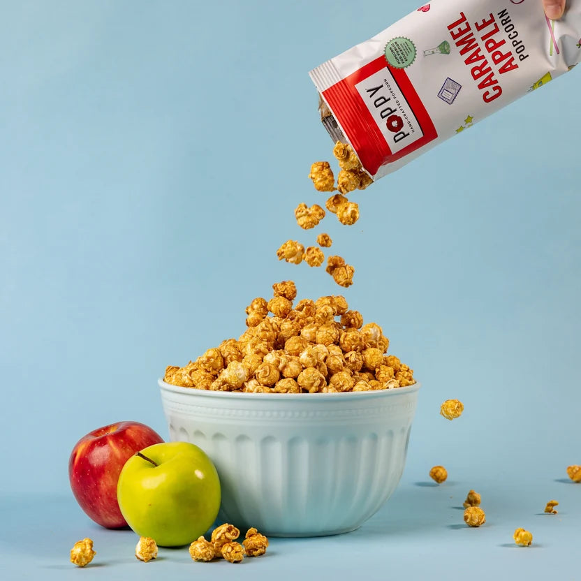 Poppy Caramel Apple Hand-Crafted Popcorn being poured into a white bowl beside fresh apples.