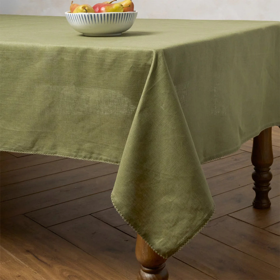 Sage green linen tablecloth with crochet edging draped over a wood dining table for an elegant everyday setting