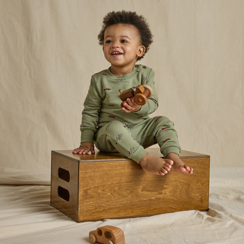 Toddler boy wearing a matching olive green race car jogger set with checkered flag print while holding a wooden toy vehicle.