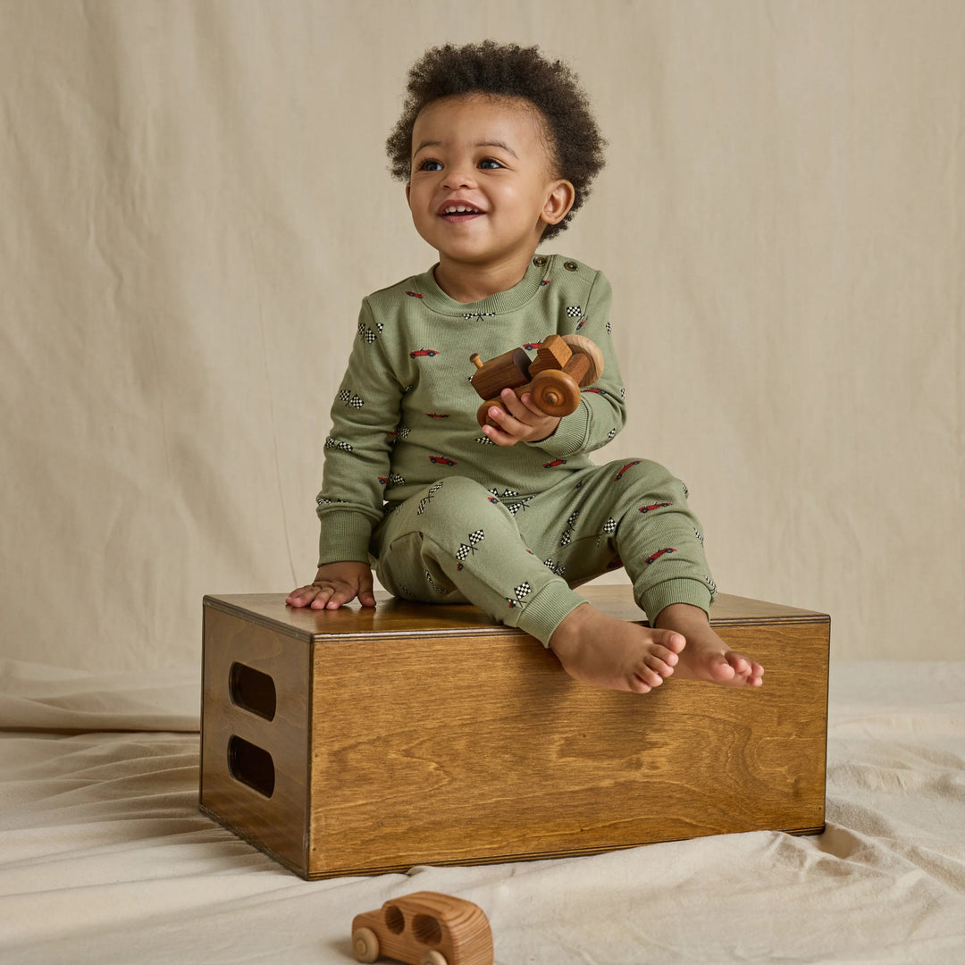 Toddler boy wearing a matching olive green race car jogger set with checkered flag print while holding a wooden toy vehicle.