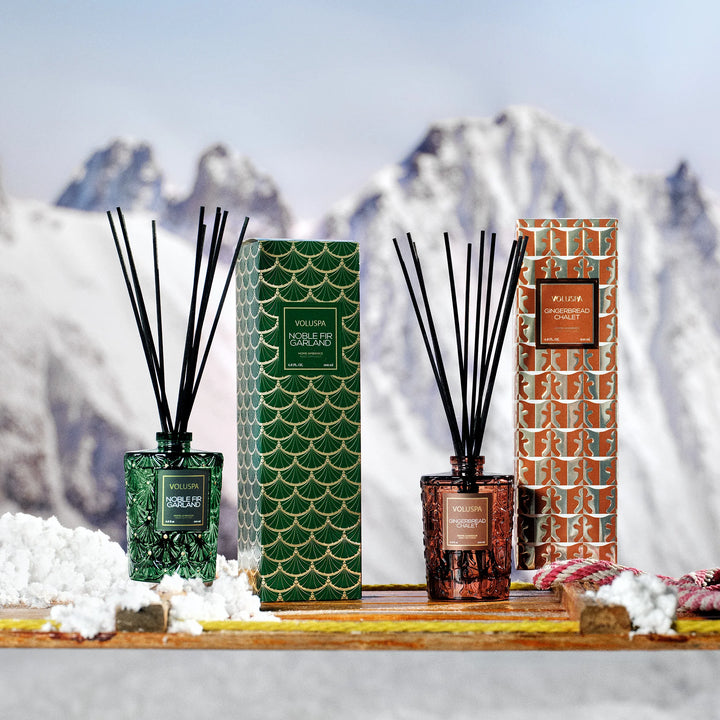 Voluspa Gingerbread Chalet Reed Diffuser displayed with Noble Fir Garland Reed Diffuser in a snowy holiday setting