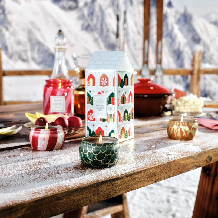 Voluspa Holiday Mini Tin Candle Trio with festive candles lit on a cozy snow-covered holiday table