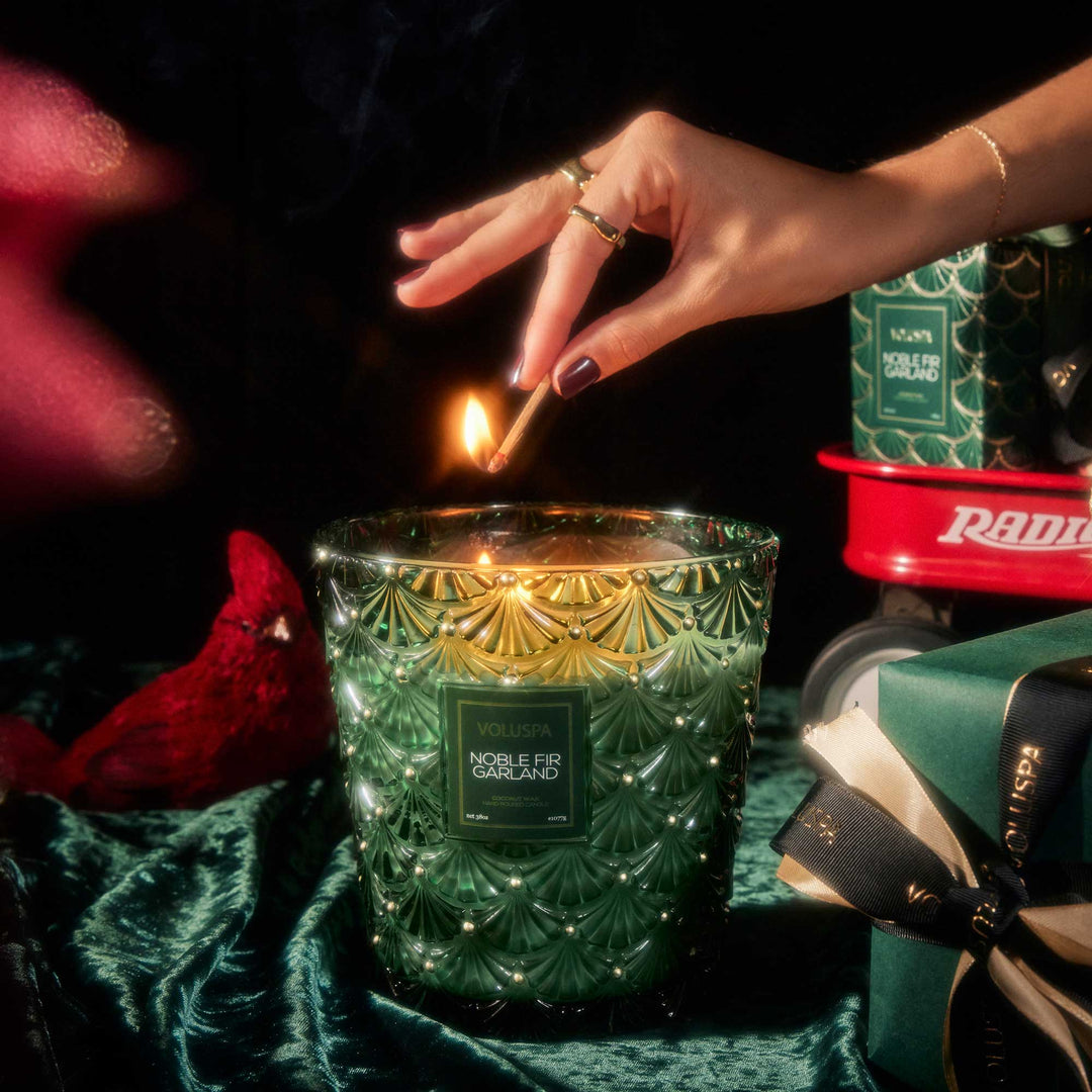 Noble Fir Garland 3 Wick Hearth Candle by Voluspa being lit beside elegant green Christmas gifts