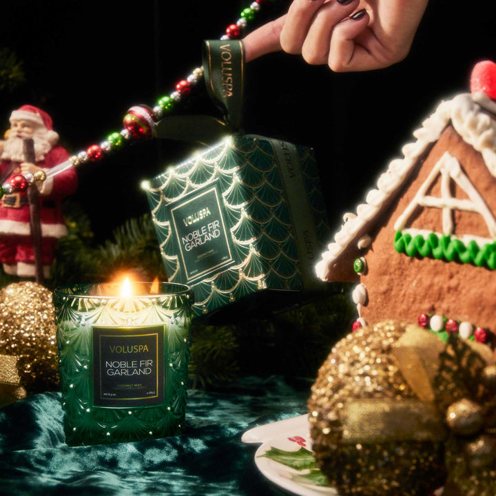 Voluspa Noble Fir Garland Classic Candle burning beside festive Christmas decorations and gingerbread house
