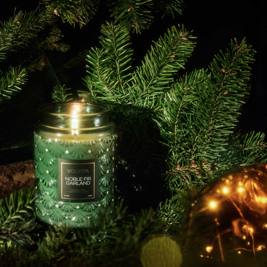 Voluspa Noble Fir Garland Large Jar Candle surrounded by fresh evergreen branches and glowing holiday lights
