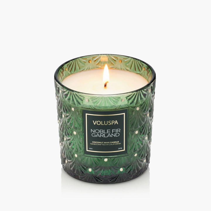 Lit Voluspa Noble Fir Garland Luxe Classic Candle in embossed emerald green glass with gold accents
