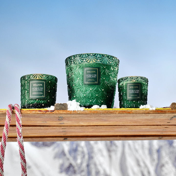 Voluspa Noble Fir Garland Luxe Classic Candle displayed with matching green holiday candles in a snowy winter scene