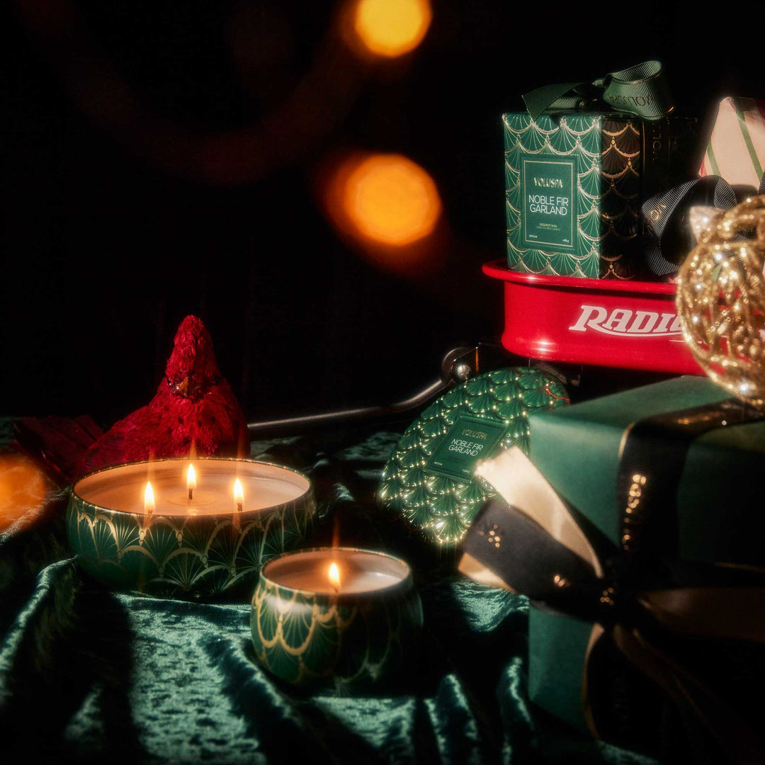Voluspa Noble Fir Garland Mini Tin Candle styled with green Christmas gifts and glowing holiday candles