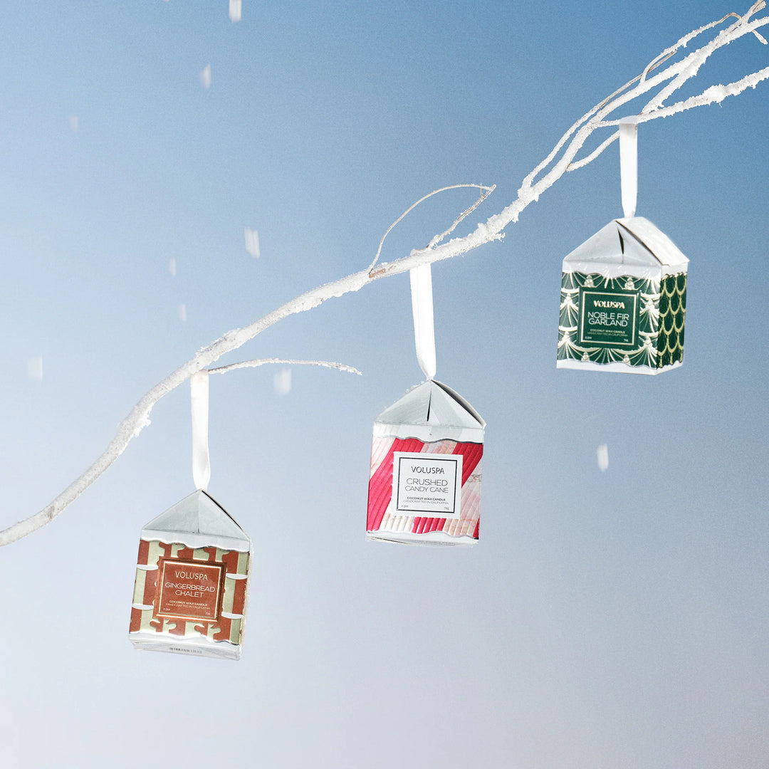 Voluspa Noble Fir Garland Ornament Pedestal Candle hanging from a snowy branch with other Voluspa holiday candle ornaments