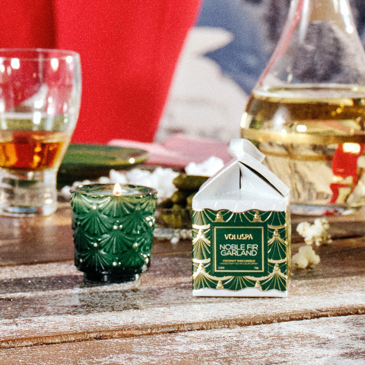 Voluspa Noble Fir Garland Ornament Pedestal Candle lit on a festive holiday table with green and red Christmas decor