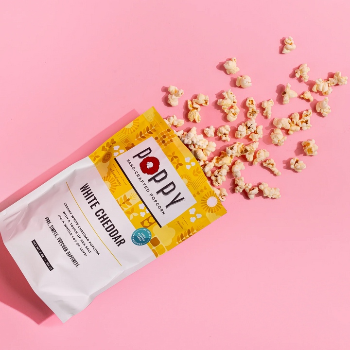 Poppy White Cheddar Hand-Crafted Popcorn bag with popcorn scattered on a pink background