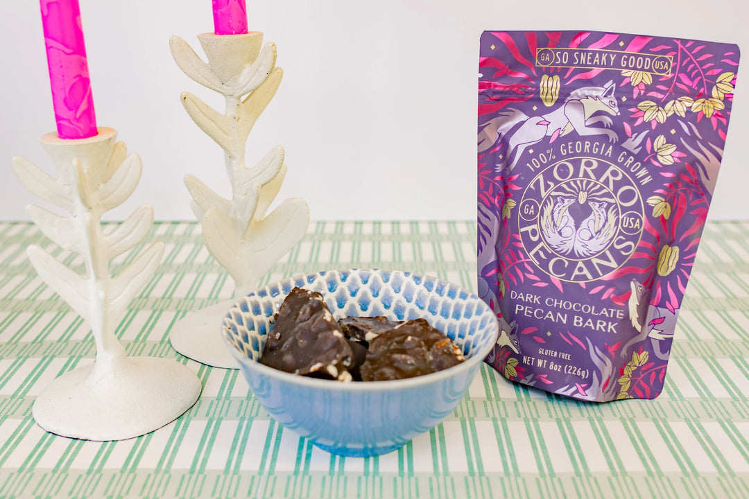 Zorro Dark Chocolate Pecan Bark in purple packaging styled with a bowl of chocolate bark and decorative pink candles.