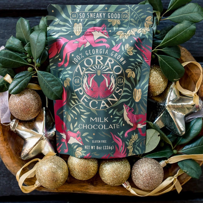 Zorro Milk Chocolate Covered Pecans in an 8 oz decorative bag surrounded by gold Christmas ornaments, greenery, and festive holiday decorations.