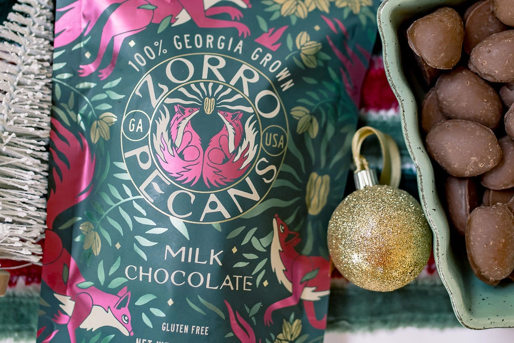 Close-up of Zorro Milk Chocolate Covered Pecans packaging beside a bowl of chocolate covered Georgia-grown pecans and a gold ornament.
